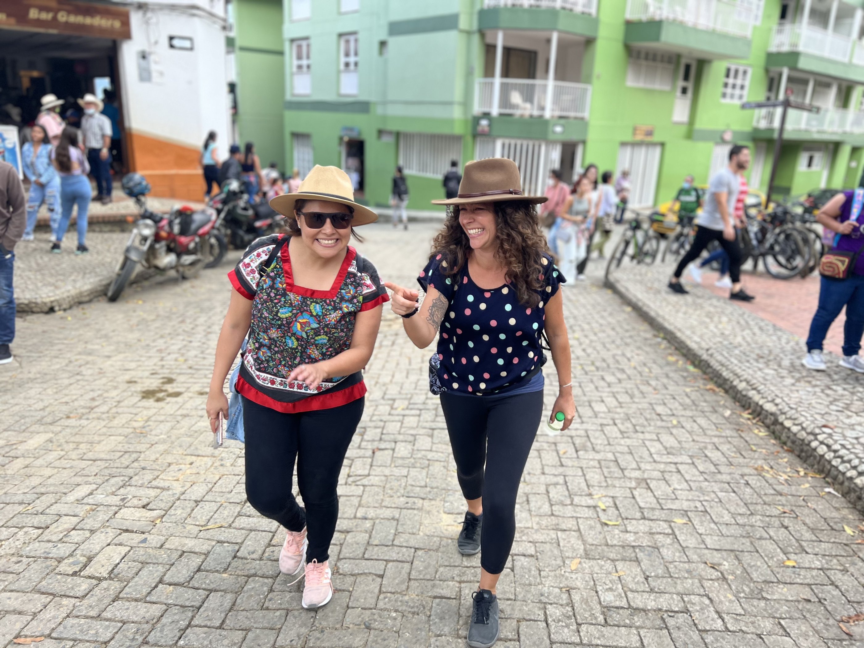 Two woman with hats on walking through the streets of Colombia. Green building in the background