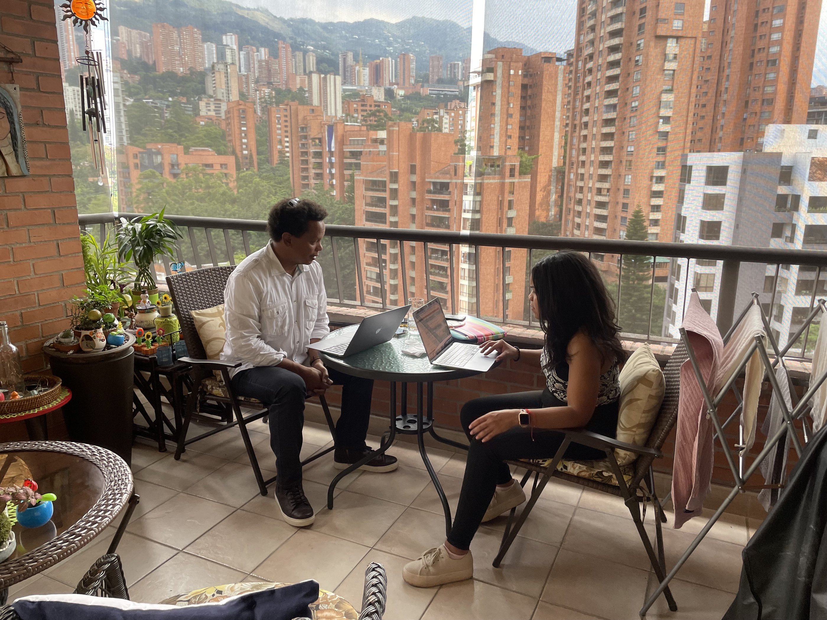 Two people sitting a bistro like table on a balcony overlooking city buildings. Mountainous views in the background 
