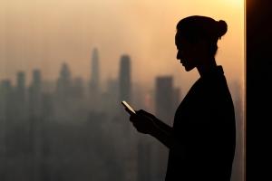 Woman working on her mobile phone next to an office window overlooking a city skyline