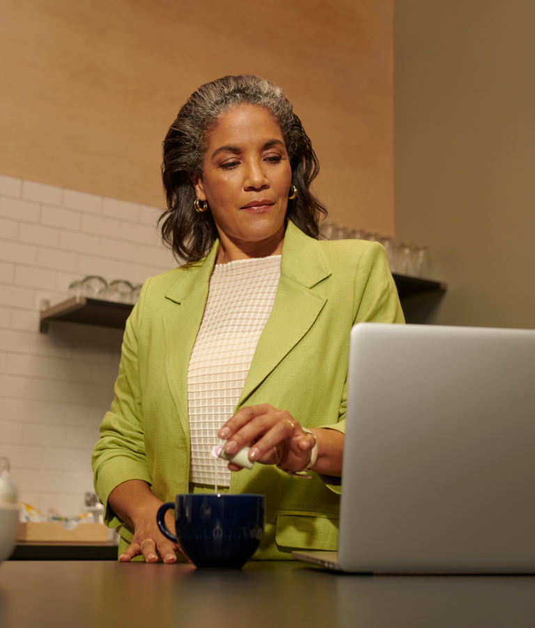 Woman in kitchen looking at laptop while brewing a cup of tea.
