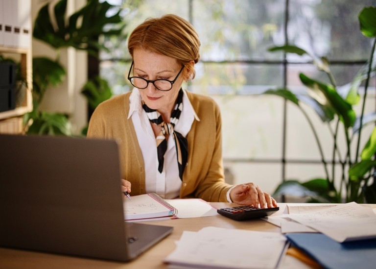 HR leader calculating payroll taxes with a calculator and logbook while sitting at her office desk