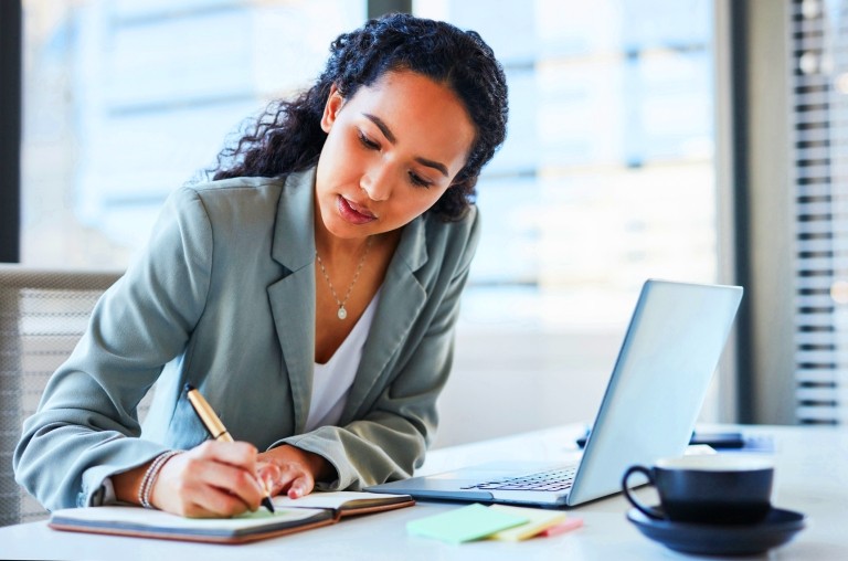 Business woman writing in a note pad with laptop and mug on desk