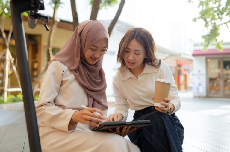 Two women sitting outside among a courtyard looking at and speaking about something shown on a tablet